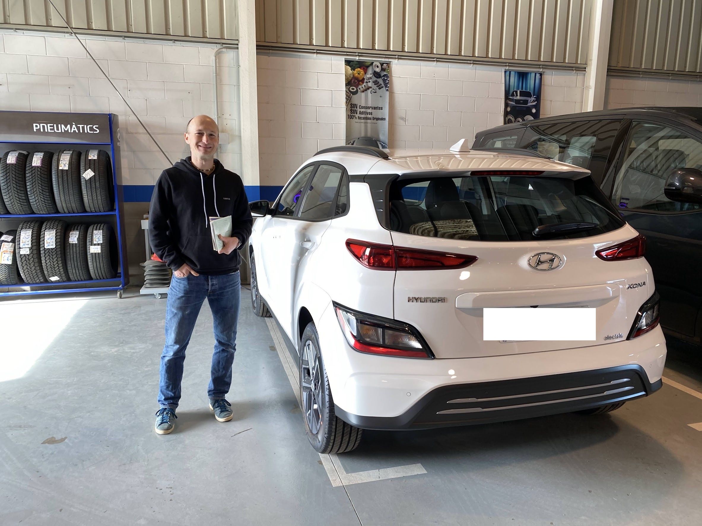 Tom in jeans and a black hoodie with the paperwork in his hands standing next to a brand-new white 2021 Hyundai Kona Electric at the car dealer.