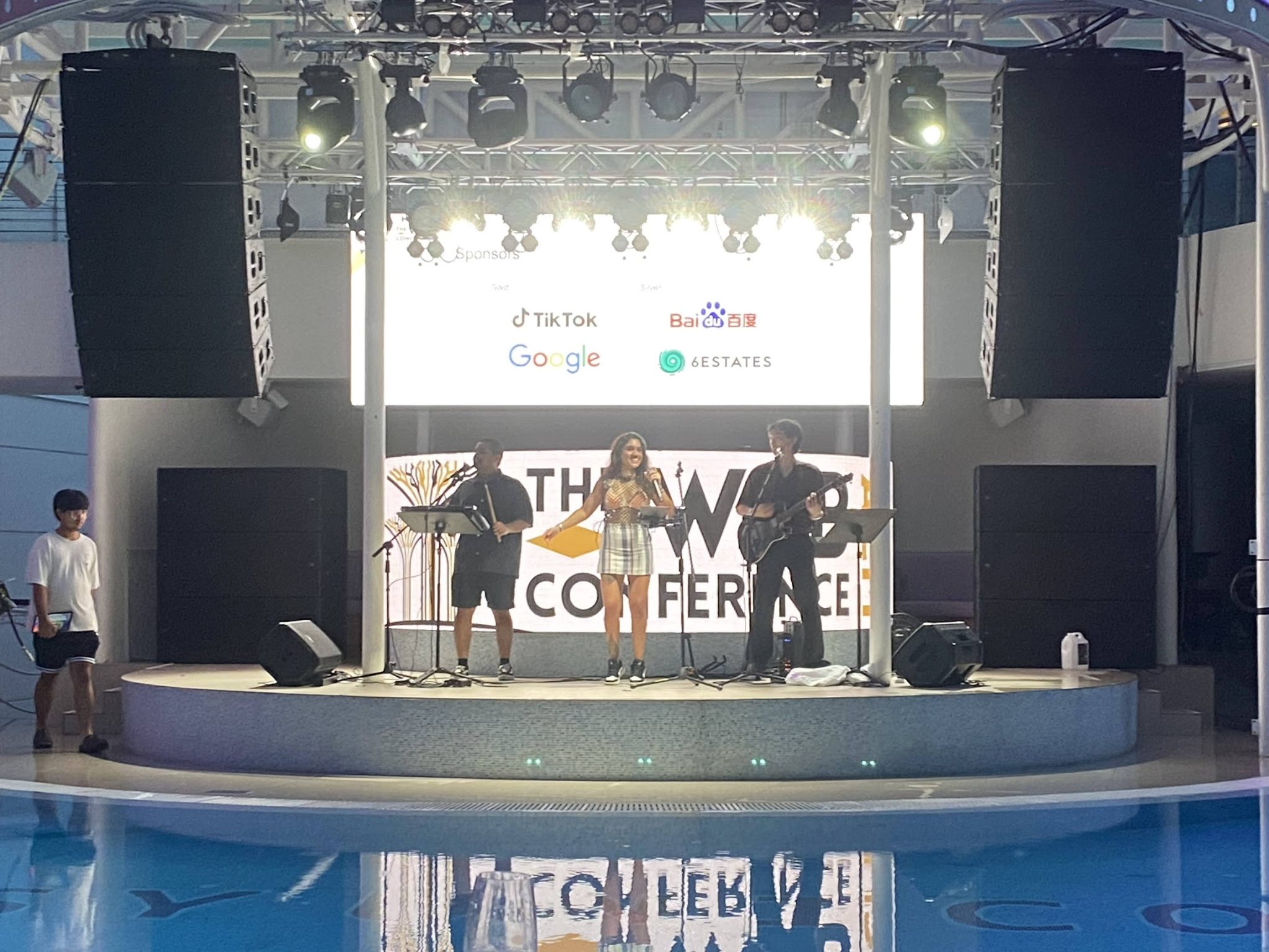 A band playing on a stage. In front of the stage is a pool. Behind the stage is a screen with a conference slide announcing the sponsors.